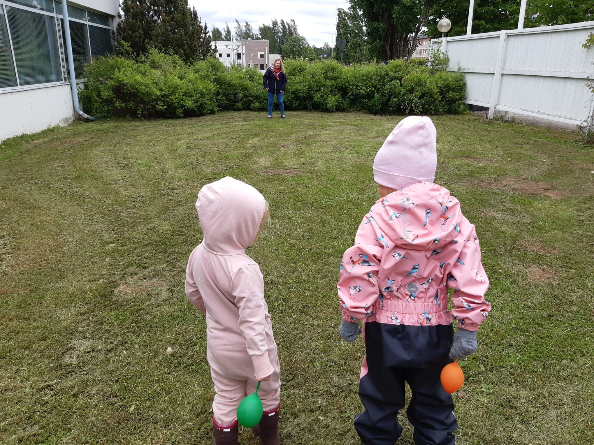 kaksi vaaleanpuna-asuista lasta etualalla, takana lastenohjaaja. yhteinen leikki menossa ulkona.