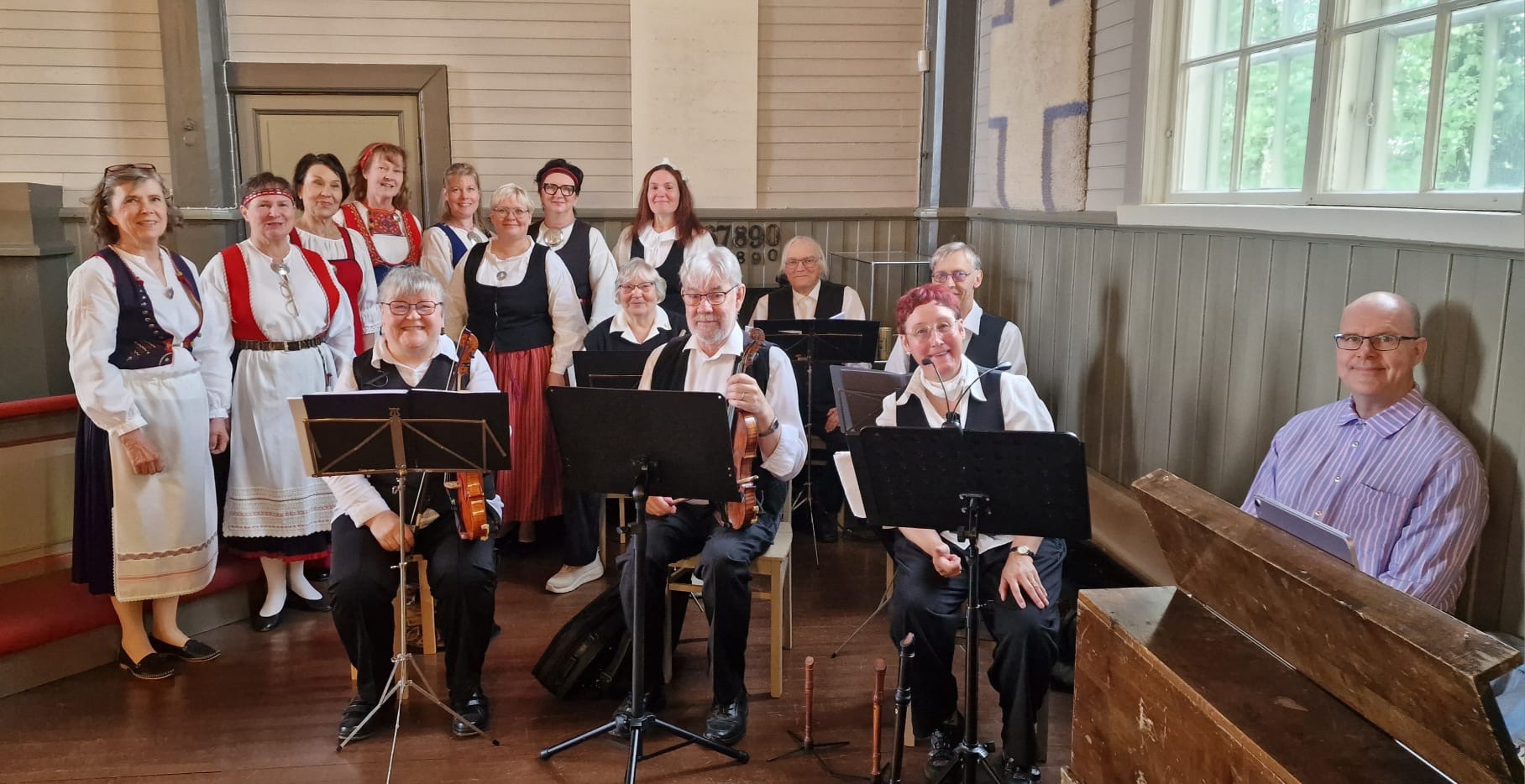 Kansallispukuihin pukeutuneita laulajia ja soittajia ryhmäkuvassa vanhassa salissa. Etualalla viulisteja nuottitelineineen, oikealla pianisti urkuharmonin ääressä.